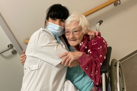Teamomat: Kollegin in weißer Dienstkleidung umarmt ältere Dame im pinken Outfit